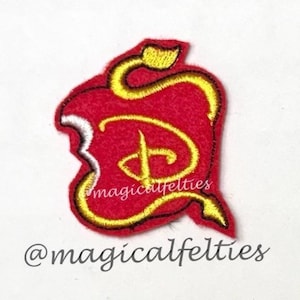 May include: A red felt patch shaped like a bitten apple with a yellow embroidered outline and a central yellow embroidered letter D. The text @magicalfelties is visible in the center of the apple and below the patch on the white surface.