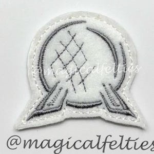 May include: A white felt patch featuring a black embroidered design of a sphere with a geometric pattern and two stylized wings. The text "@magicalfelties" is below the patch.