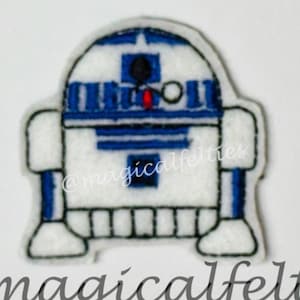 May include: A white and blue felt applique of R2-D2, a robot character from the Star Wars franchise. The applique is embroidered with details and has a white background.