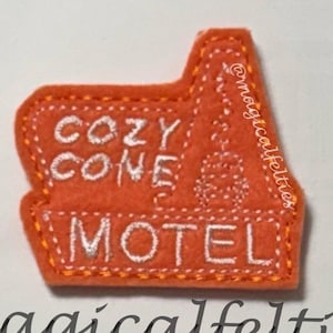 May include: An orange felt motel-shaped patch with white embroidered text that reads "COZY CONE MOTEL". The patch has a stitched border and a small design on the right side. The text "@magicalfelties" is written below the patch.