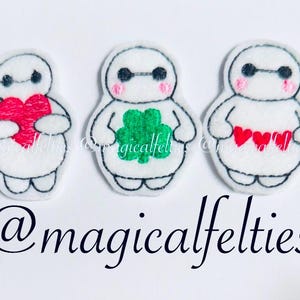 May include: Three white felt appliques of a robot character holding a red heart, a green four-leaf clover, and three red hearts. The appliques are embroidered with black outlines and pink cheeks. The text "@magicalfelties" is at the bottom of the image.