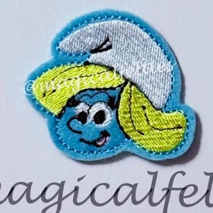 May include: A blue felt patch featuring a cartoon character with a white hat and yellow hair. The character has a blue face, black eyes, and a smiling mouth. The patch is outlined in blue and has the text "magicalfelti" at the bottom.