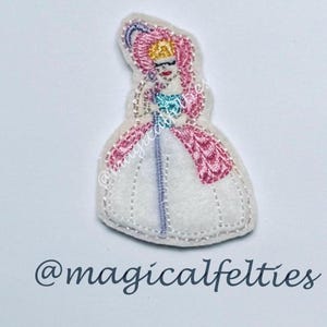 May include: Embroidered felt princess doll with pink hair, a white dress, and a blue and purple staff. The doll has a gold crown and red lips. The text "@magicalfelties" is at the bottom.