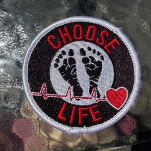 May include: A circular embroidered patch with a black background and a white border. The text CHOOSE LIFE is stitched in red. In the center, two black baby footprints are depicted above a red EKG heart rhythm line ending in a red heart.