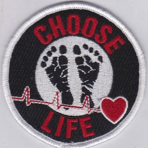Puede incluir: Un parche bordado en blanco y negro con un círculo blanco en el centro. El círculo tiene dos huellas de pies negras en su interior. Las palabras "CHOOSE LIFE" están en rojo alrededor del círculo. Un corazón rojo y una línea de latido del corazón roja están debajo del círculo.