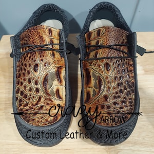 May include: A pair of black slip-on shoes with a brown alligator print leather upper. The shoes have a black elastic band across the top and a black rubber sole. The text "crazy arrow custom leather & more" is printed on the bottom of the shoes.