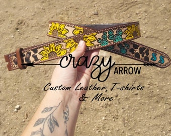 May include: A brown leather belt with a sunflower and cactus design. The belt buckle is silver and has the words "Crazy Arrow" engraved on it. The belt is decorated with black and white spots and the initials "SS" are on the end of the belt.