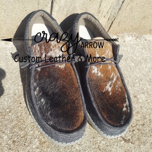 May include: A pair of brown and white cowhide slip-on shoes with black canvas trim. The shoes have a  "crazy arrow custom leather & more" logo on the side.