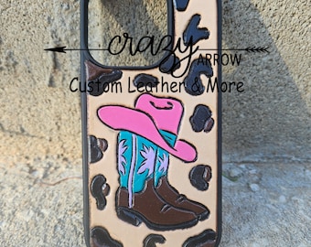 May include: A brown leather phone case with a pink cowboy hat and brown cowboy boots on a tan and brown leopard print background. The text "crazy arrow custom leather & more" is printed on the case.