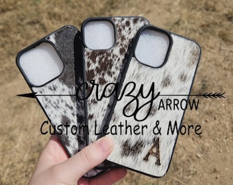 May include: Three phone cases with cowhide patterns in black, brown, and white are displayed. The cases have black borders around the camera openings. The text "Crazy Arrow Custom Leather & More" is visible on the cases.