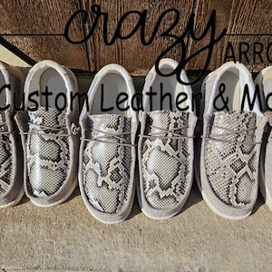 May include: Five pairs of grey suede slip-on shoes with a snakeskin pattern. The shoes are arranged in a row and have a white sole. The text "Crazy Arrow Custom Leather & More" is visible above the shoes.