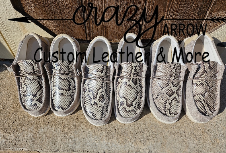 May include: Five pairs of grey suede slip-on shoes with a snakeskin pattern. The shoes are arranged in a row and have a white sole. The text "Crazy Arrow Custom Leather & More" is visible above the shoes.