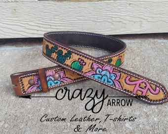 May include: A brown leather belt with a tan background and colourful painted cactus and flower designs. The belt buckle is brown with a silver accent. The belt is labelled "crazy arrow" and "Custom Leather, T-shirts & More".