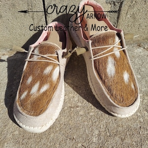 May include: A pair of brown and white cowhide slip-on shoes with pink lining. The shoes have a white sole and are tied with a beige shoelace. The text "crazy arrow custom leather & more" is visible on the shoes.