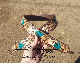 May include: Brown leather strap with a tooled design of white feathers and teal accents. The strap is decorated with a floral pattern and has a buckle on one end. The strap is labeled "crazy arrow custom leather, t-shirts & more".