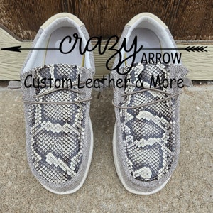 May include: A pair of grey canvas slip-on shoes with a snake print design. The shoes have a white sole and a brown leather lace. The text "crazy arrow custom leather & more" is visible on the shoes.