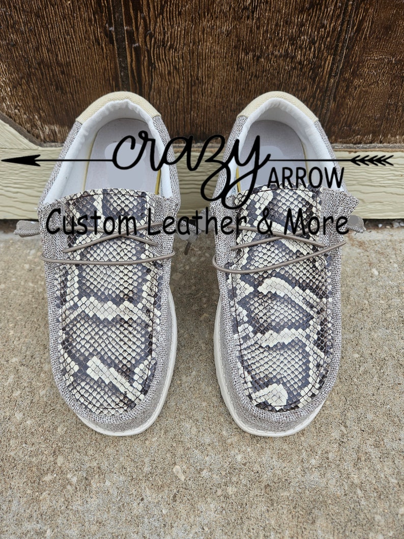 May include: A pair of grey canvas slip-on shoes with a snake print design. The shoes have a white sole and a brown leather lace. The text "crazy arrow custom leather & more" is visible on the shoes.