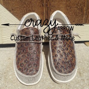 May include: A pair of brown leather slip-on shoes with a beige canvas trim. The shoes have a crocodile embossed pattern and the text "crazy arrow custom leather & more" is visible on the shoes.