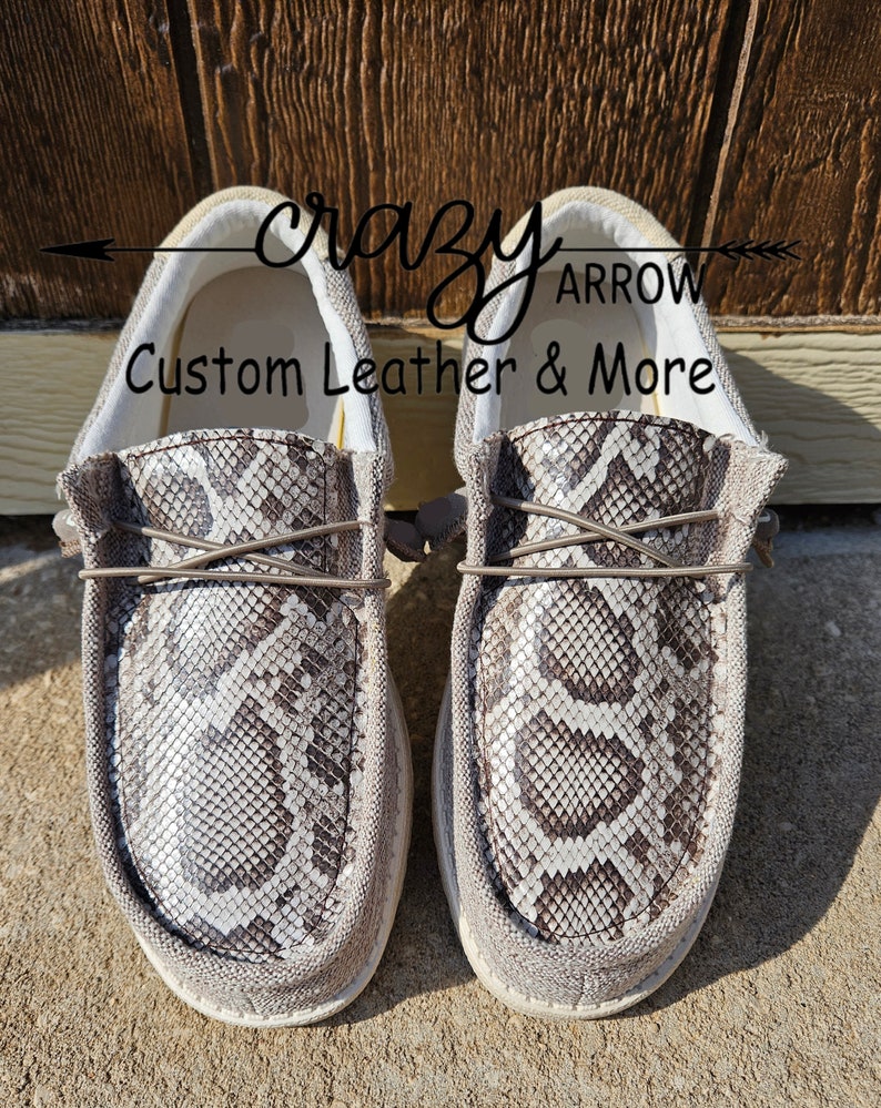 May include: A pair of grey canvas slip-on shoes with a snakeskin pattern. The shoes have a white sole and a brown leather lace. The text "crazy arrow custom leather & more" is printed on the shoes.