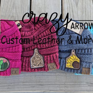 May include: Four different colored knit beanies with various leather patches. The beanies are pink, burgundy, gray, and blue. The patches include a cactus, a floral design, a sunflower, and a cow.