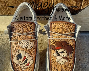 May include: A pair of gray canvas shoes with custom tooled leather panels. One shoe features a skull with a cowboy hat and a cigar, while the other shoe features playing cards and dice. The shoes are labeled "Crazy Arrow Custom Leather & More."