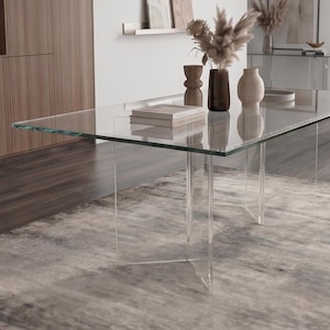 May include: A modern dining room featuring a large, rectangular glass table with clear acrylic legs. The table is set on a patterned rug. Decorative vases and a plant in a beige pot add to the room's decor. A modern light fixture hangs above.