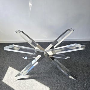 May include: A clear acrylic table with a starburst design. The table has multiple angular legs that extend outwards from a central point. The legs are approximately 20 inches long. The table is on a gray carpet.
