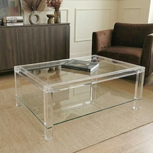 May include: A modern living room featuring a clear acrylic coffee table with a lower shelf, a brown sofa, and a matching armchair. A neutral rug and a dark wood cabinet with decor complete the scene. A book rests on the table.