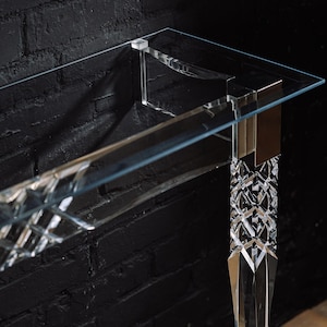 Könnte beinhalten: Ein durchsichtiger Glaskonsolentisch mit geometrischem Design. Der Tisch hat eine rechteckige Tischplatte und zwei Beine mit einem facettierten Design.