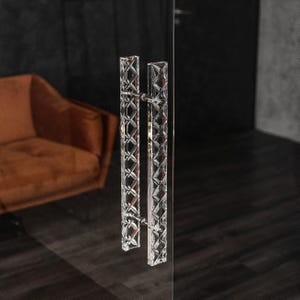 Op de afbeelding: Een paar heldere glazen deurgrepen met een geometrisch patroon. De grepen zijn bevestigd aan een glazen deur.