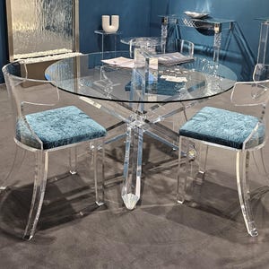May include: A contemporary dining set with a clear glass tabletop and acrylic legs. Two acrylic chairs with teal velvet seat cushions are around the table. The table's base has a starburst design. The set is in a room with a dark blue wall.