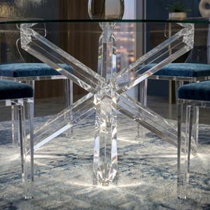 May include: A modern dining table with a clear glass top and a unique, geometric base made of clear acrylic. The table is surrounded by chairs with blue velvet seats. The table's base has an X-shaped design. The table is set on a patterned rug.