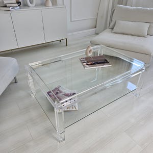 Peut inclure: Une table basse en acrylique transparent avec un plateau rectangulaire et une étagère inférieure. La table a un design moderne avec des lignes droites et est soutenue par quatre pieds transparents. Des magazines et un vase décoratif sont sur la table.