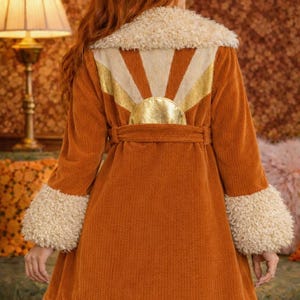 May include: A rust-coloured corduroy coat with a cream-coloured faux fur collar and cuffs. The back features a sunburst design with gold accents. The coat has a matching belt and a flared skirt.
