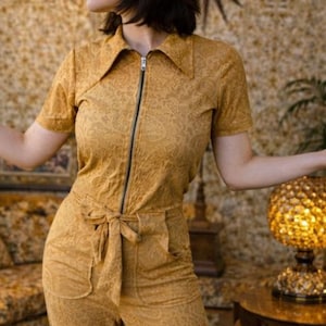 May include: Mustard yellow jumpsuit with a paisley pattern. The short-sleeved jumpsuit features a collared neckline, a front zip, and a tie belt at the waist. The jumpsuit has two front pockets and wide-leg trousers. The model is wearing black boots.