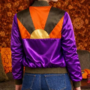 Może przedstawiać: Fioletowa satynowa kurtka bomberka z pomarańczowym i złotym wzorem wschodu słońca na plecach. Kurtka ma zielony kołnierz i mankiety. Osoba na zdjęciu ma na sobie niebieskie dżinsy.