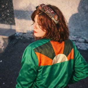 Può includere: Giacca di raso verde con dettagli arancioni e neri sul retro, con un design a raggiera. La giacca ha un cerchio bianco al centro del design. Viene indossata una fascia per capelli fantasia.