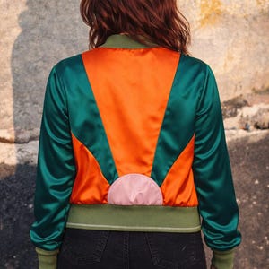 Può includere: Giacca bomber in raso con un design accattivante. La parte posteriore presenta un motivo a forma di sole in arancione, verde e rosa. La giacca ha maniche verdi e orlo e polsini a coste verdi. La modella indossa jeans neri.