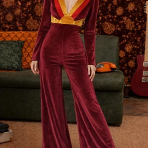 Könnte beinhalten: Burgunderfarbener Samt-Jumpsuit mit langen Ärmeln und ausgestellten Beinen. Der Ausschnitt hat eine V-Form mit einem gold-, rot- und gelbfarbenen Design. Der Jumpsuit ist im Retro-Stil gehalten und eignet sich für Partys und besondere Anlässe.
