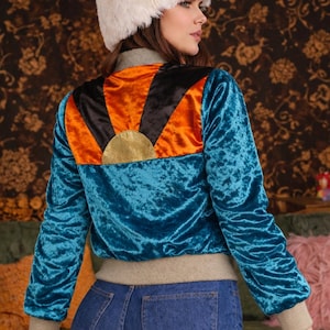 Könnte beinhalten: Türkisblaue Samt-Bomberjacke mit einem orange-schwarzen Sonnenmuster auf dem Rücken. Die Jacke hat ein goldenes Sonnendetail und einen beigefarbenen gerippten Kragen und Bündchen. Das Model trägt eine blaue Jeans und einen weißen Pelzhut.