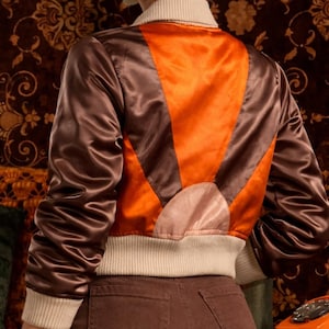 May include: A brown and orange satin bomber jacket with a cream ribbed collar, cuffs, and hem. The back of the jacket features a sunburst design in orange and brown. The jacket is worn over brown trousers.