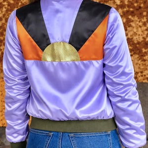 Op de afbeelding: Een lavendelkleurige satijnen bomberjack met een opvallend geometrisch ontwerp in zwart, oranje en goud op de rug. Het heeft een groene geribbelde kraag, manchetten en zoom. Het model draagt een blauwe jeans. Een stijlvolle jas.