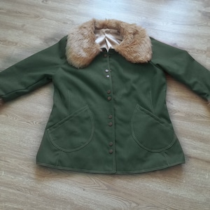 Op de afbeelding: Een groene jas met een bruine nep bontkraag en manchetten. De jas heeft een knoopsluiting en twee grote zakken.