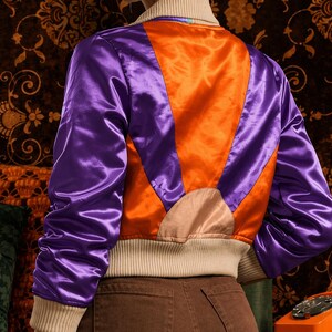 Könnte beinhalten: Satin-Bomberjacke in Lila und Orange. Die Jacke hat einen cremefarbenen gerippten Kragen und Bündchen. Die Rückseite der Jacke hat ein Sonnenstrahl-Design in Orange und Lila.