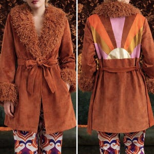 Peut inclure: Un long manteau de couleur rouille avec un col et des poignets en fausse fourrure. Le manteau est doté d'une ceinture à nouer et d'un motif de soleil coloré au dos. Le manteau est associé à un pantalon à motifs, créant une esthétique rétro et bohème.