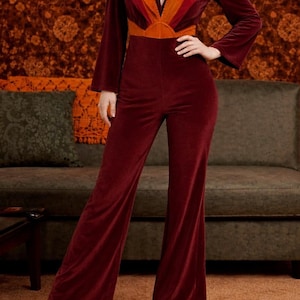 Könnte beinhalten: Burgunderfarbener Samt-Jumpsuit mit tiefem Ausschnitt und ausgestellten Beinen. Das Oberteil hat ein Colorblock-Design in Orange- und Rottönen. Die langen Ärmel und weiten Beine erzeugen einen Retro-Look im 70er-Jahre-Stil.