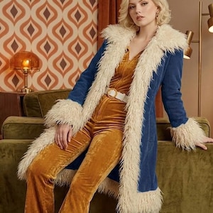 Può includere: Un lungo cappotto in velluto a coste blu con un bordo spesso in pelliccia sintetica color crema lungo i revers e i polsini. Il cappotto è indossato sopra una tuta in velluto color senape con una cintura bianca con fibbia circolare.