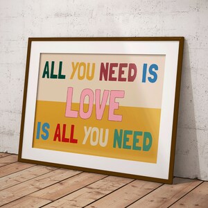 A framed Beatles print leans against a wooden floor. The music poster features a split yellow and beige background with the lyrics 'All You Need is Love' written is multiple colours.