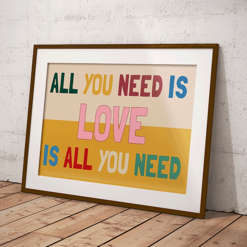 A framed Beatles print leans against a wooden floor. The music poster features a split yellow and beige background with the lyrics 'All You Need is Love' written is multiple colours.