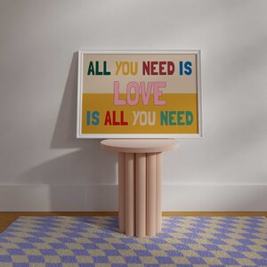 A framed Beatles music print is displayed above a checkerboard rug. The poster features the All You Need is Love song lyrics against a yellow and beige background.